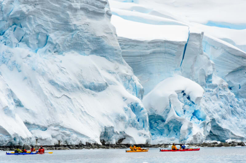 Kayakers in drysuits beneath bulging glacial ice formations at Melchior Island, western Antarctic Peninsula