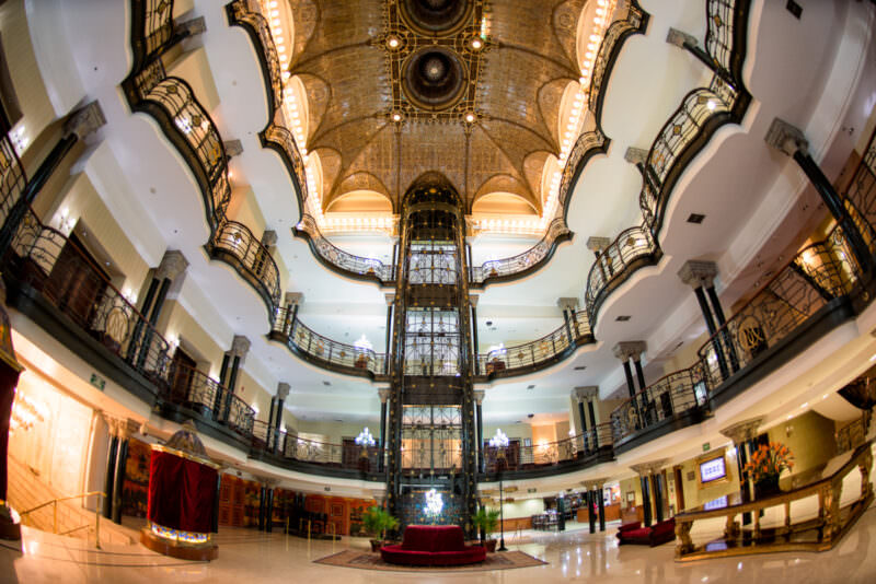 Fisheye view of Gran Hotel Ciudad de Mexico atrium with wrought-iron cage elevator rising through four floors of curved balconies