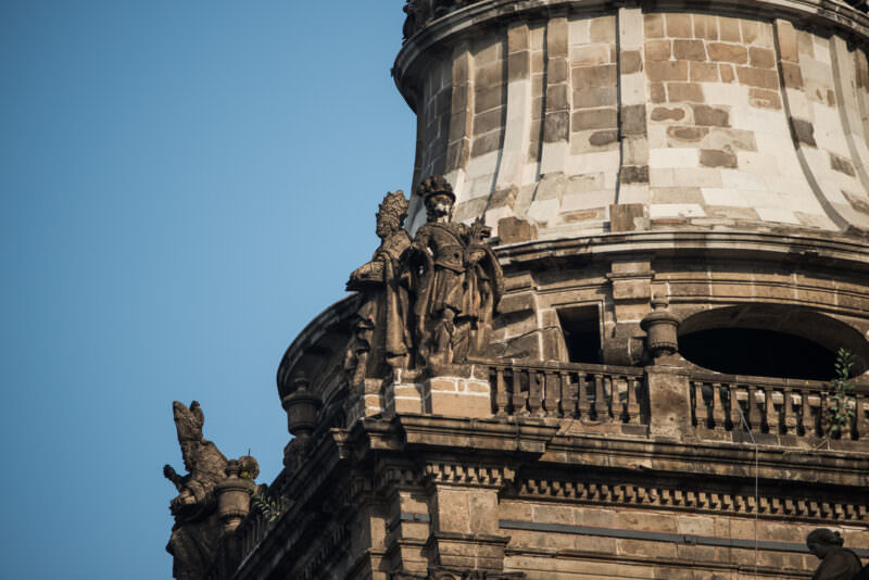 Stone saints flanking the drum of a bell tower, carved in white chiluca against the sky