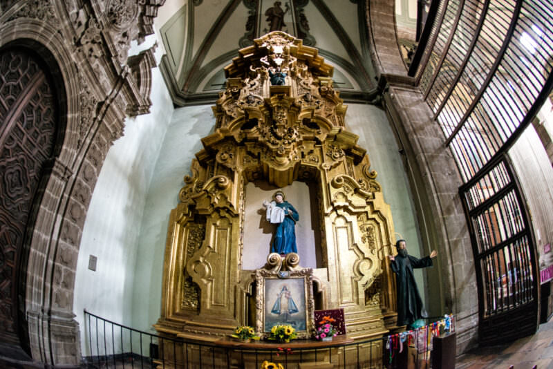 Gilded retablo in a side chapel holding a blue-robed Virgin, dark friar figure, and sunflowers at the rail