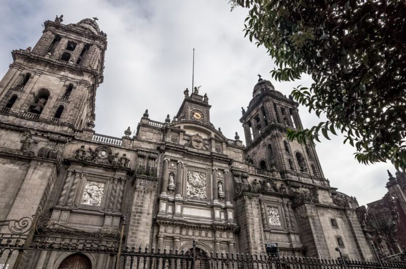 Catedral Metropolitana south facade rising above the Zócalo, bell towers and Neoclassical clock against cloudy sky