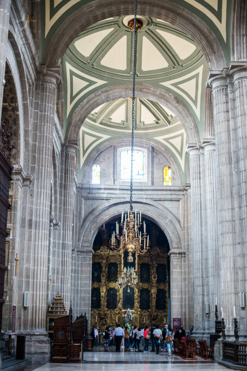 Cathedral side aisle stretching toward a gilded retablo beneath pale ribbed vaulting, visitors gathered below