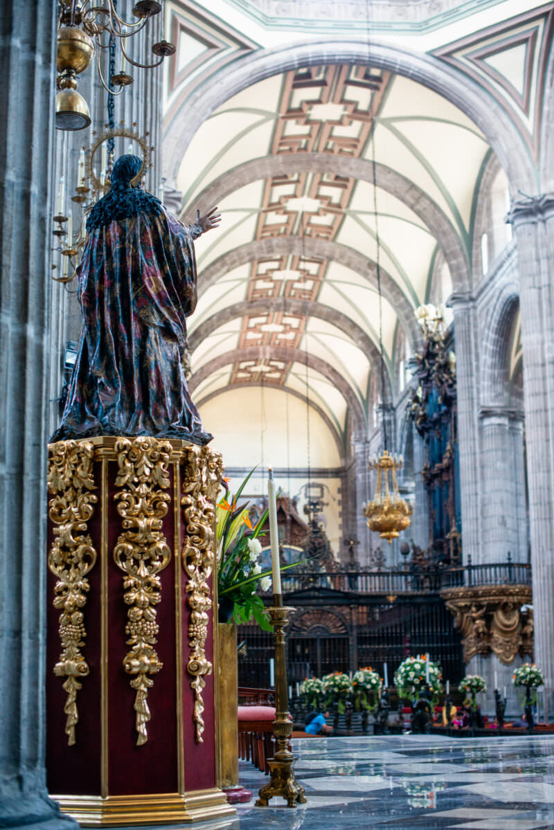 Dark-robed colonial figure on gilded pedestal facing the nave, bronze choir screen in the middle distance