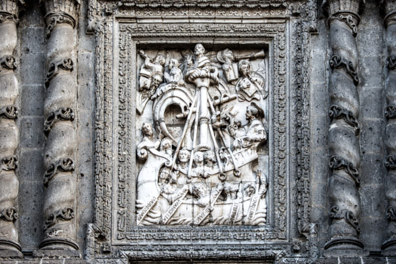 Chiluca stone relief panel on El Sagrario facade depicting a Eucharistic scene with scrolls and saints