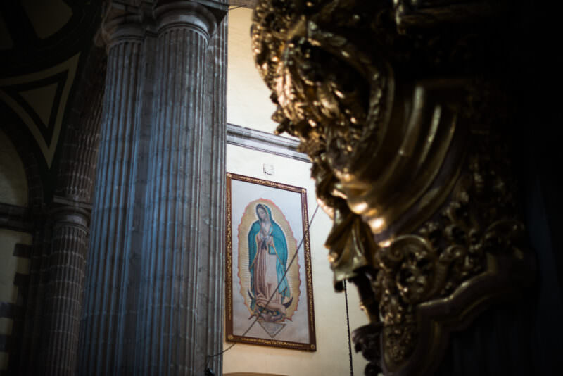 Reproduction of Our Lady of Guadalupe image hanging between a dark column and a gilded retable scroll