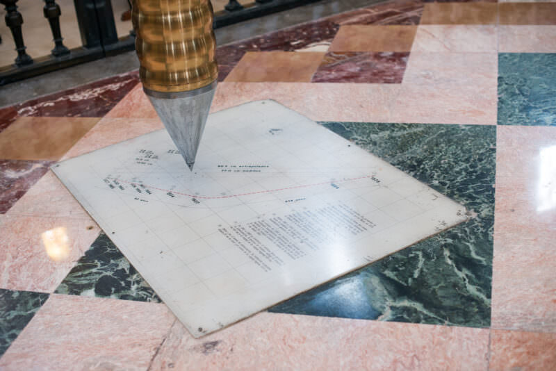 Brass pendulum tip hovering over floor-mounted settlement tracking chart with Spanish measurement text