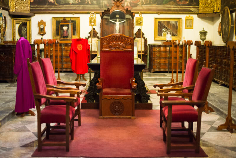 Archbishop's carved wooden throne flanked by crimson chairs in the sacristy, liturgical vestments hung on racks