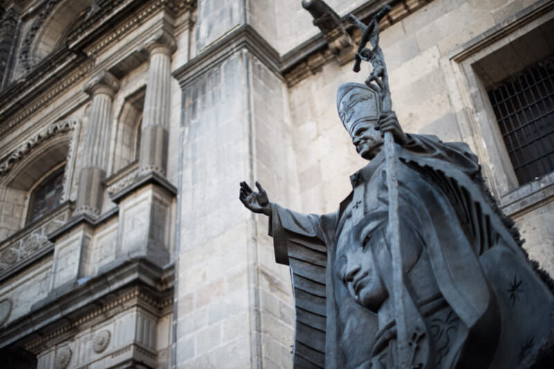 Bronze sculpture of Pope John Paul II raising his hand in blessing, seen from below against the cathedral facade