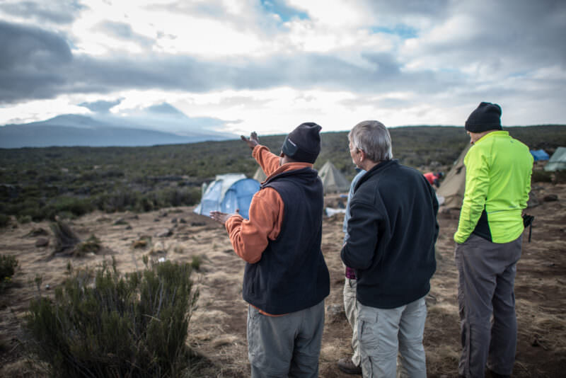 A guide gestures toward Kibo's cloud-wrapped summit dome as trekkers look on from tents at Shira 1 Camp on Kilimanjaro