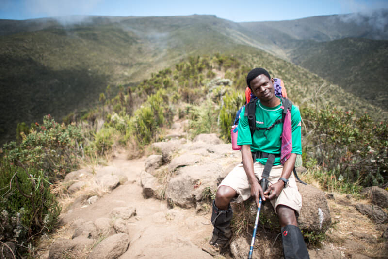 A guide resting trailside on Kilimanjaro's Lemosho Trail amid heath-zone shrubs and layered green ridges