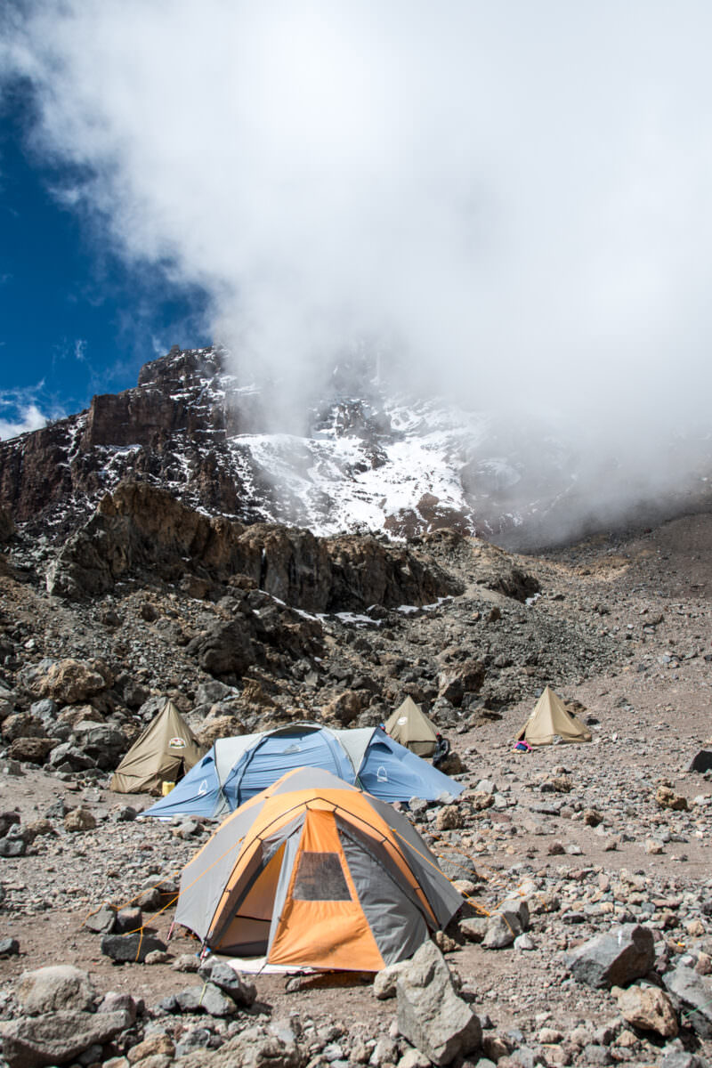 Colorful tents dot the rocky ground of Arrow Glacier Camp beneath Kilimanjaro's snow-streaked Western Breach cliffs