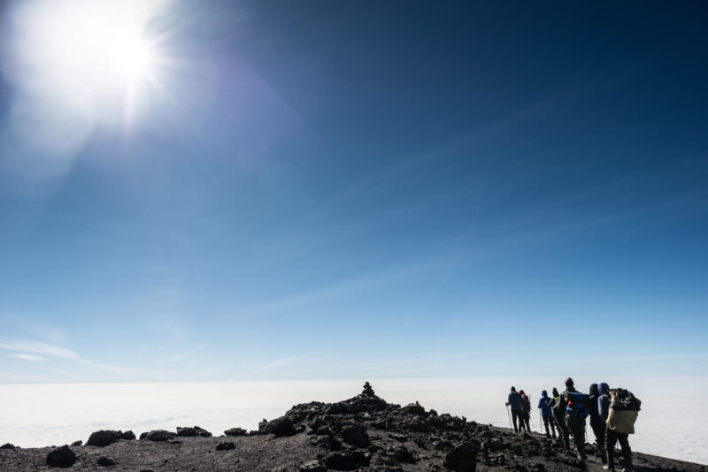 Silhouetted climbers along Kibo crater's rocky rim near Uhuru Peak with sun flare and clouds below