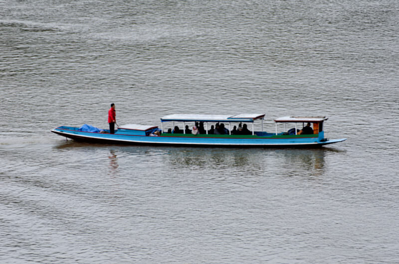 Covered blue-and-white passenger boat with passengers aboard navigating the wide Nam Ou River at Nong Khiaw