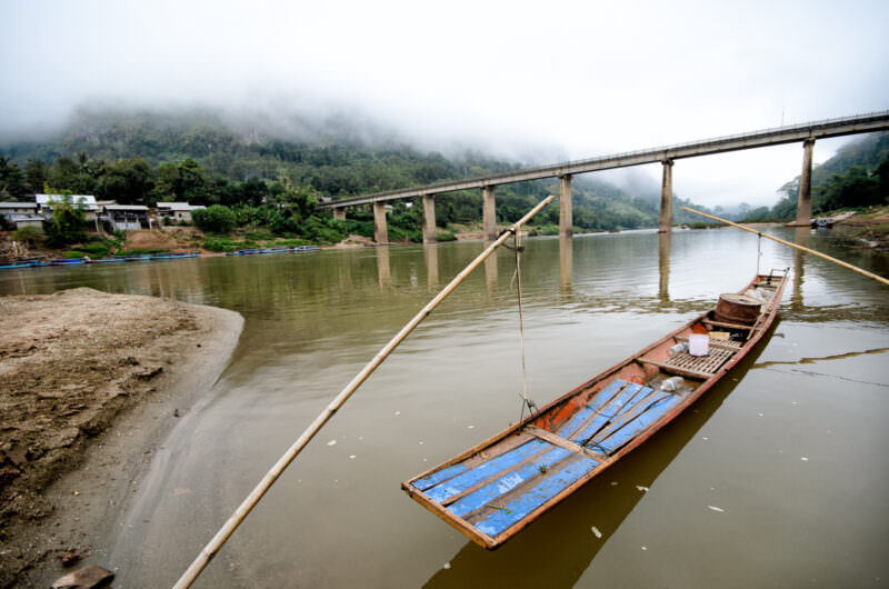 Traditional wooden longtail boat moored beneath Nong Khiaw's concrete bridge, misty karst mountains rising behind