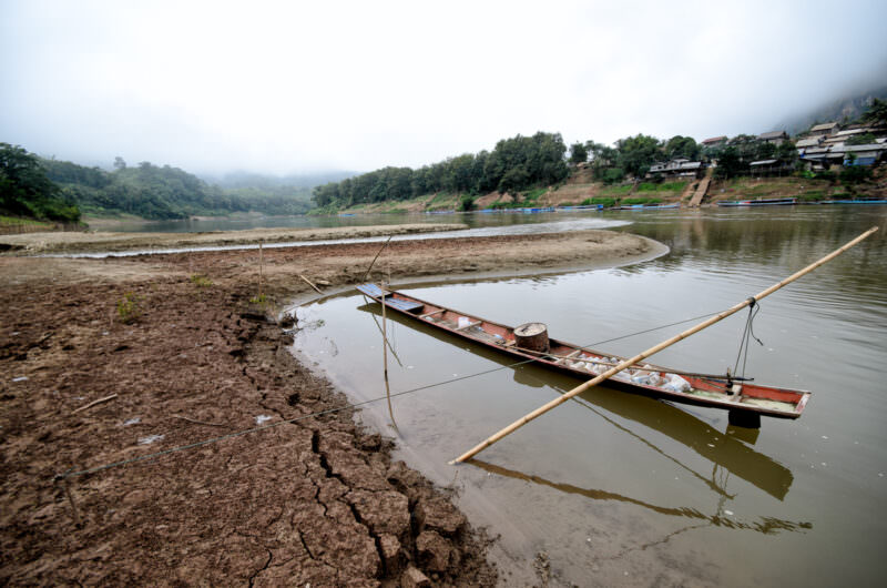 Wooden longtail boat moored against deeply cracked mudbank along the Nam Ou during low water, hillside village in the distance