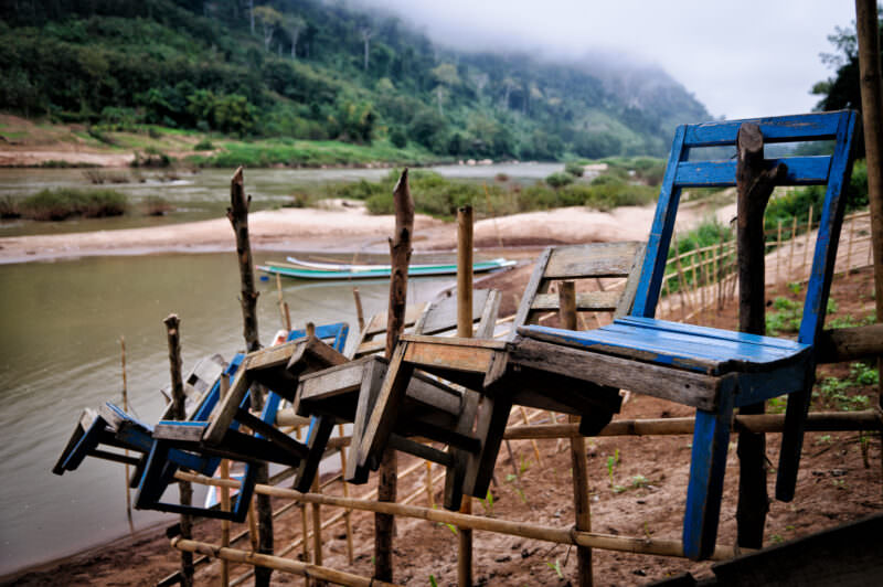 Wooden chairs hanging from a bamboo fence protecting a riverside vegetable plot, Nam Ou and mist-shrouded karst beyond