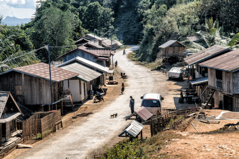Wooden stilt houses clustered along a winding dirt road carved through forested mountains, vehicles and people visible