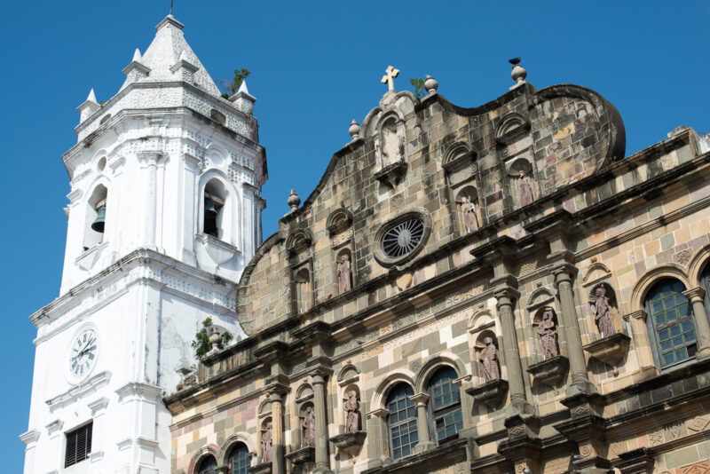 Catedral Basílica Santa María la Antigua in Panama City seen from below, its weathered carved stone facade rising between two white bell towers against a blue sky