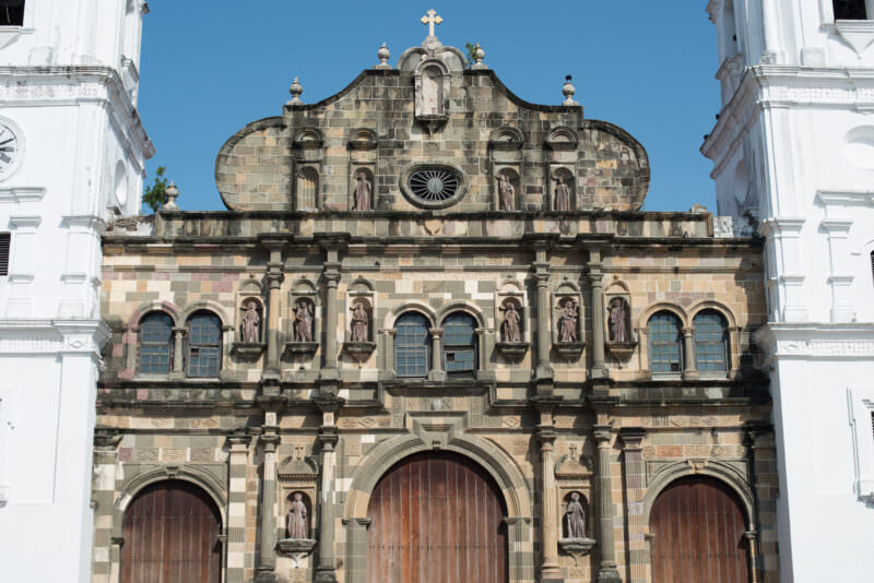 Weathered stone facade of the Catedral Metropolitana showing three tiers of carved statues, arched windows, a round oculus, and crosses atop the curved gable, flanked by white bell towers