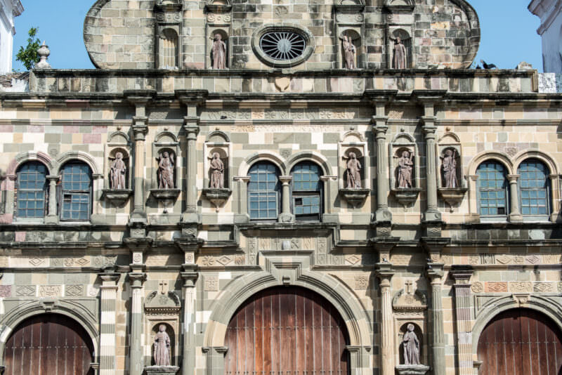 Weathered stone facade of the Catedral Metropolitana with three arched wooden doors, carved statues in niches, arched windows, and a curved gable with crosses atop