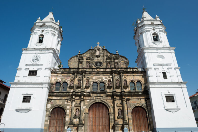 Catedral Basílica Santa María la Antigua viewed from Plaza de la Independencia under a blue sky, its weathered stone facade with carved statues between two white bell towers