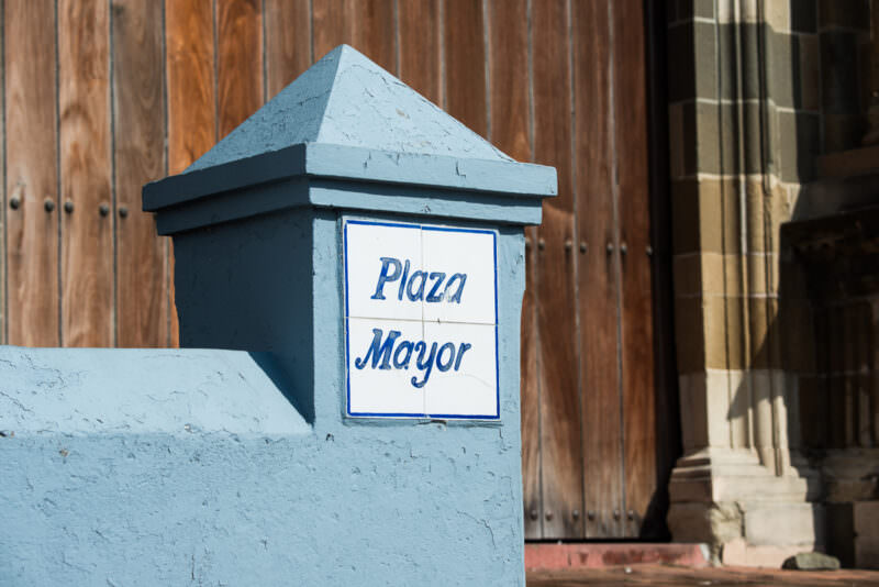 Blue-and-white ceramic tile sign reading 'Plaza Mayor' mounted on a gray post in front of wooden cathedral doors and a stone column