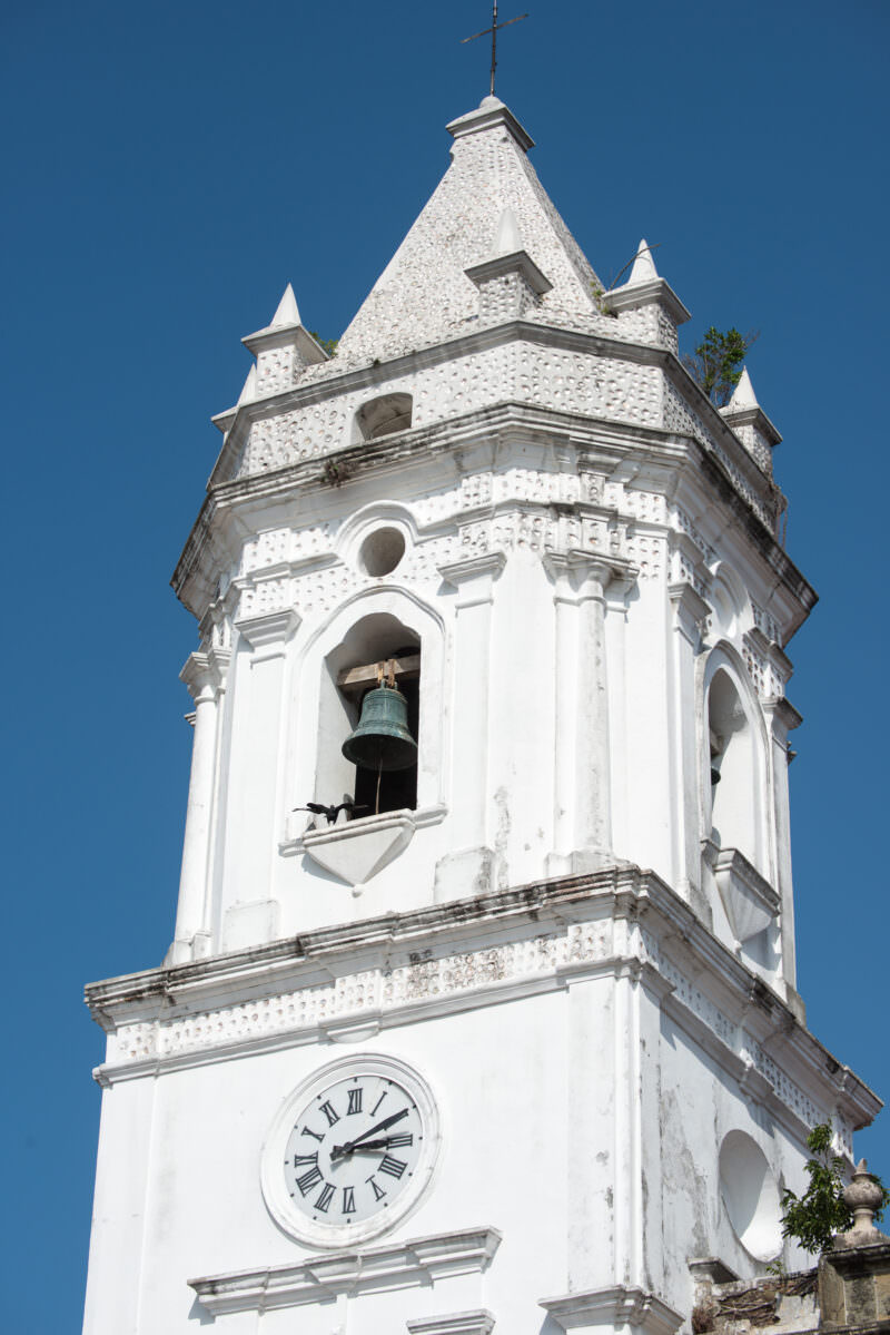 White bell tower of the Catedral Metropolitana with mother-of-pearl-inlaid pyramidal roof rising above colonial rooftops, its arched openings and bronze bell visible against blue sky