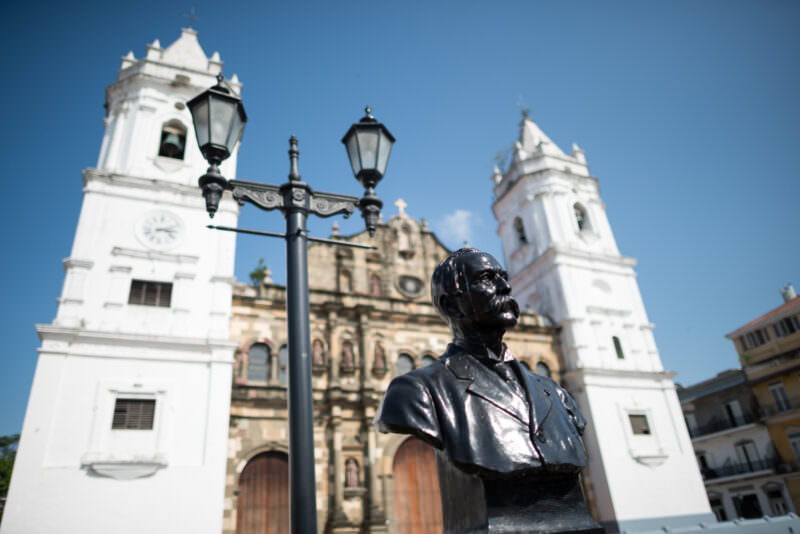 Bronze bust of Dr. Manuel Amador Guerrero in foreground with the Catedral Metropolitana facade and twin white bell towers behind, seen from Plaza de la Independencia
