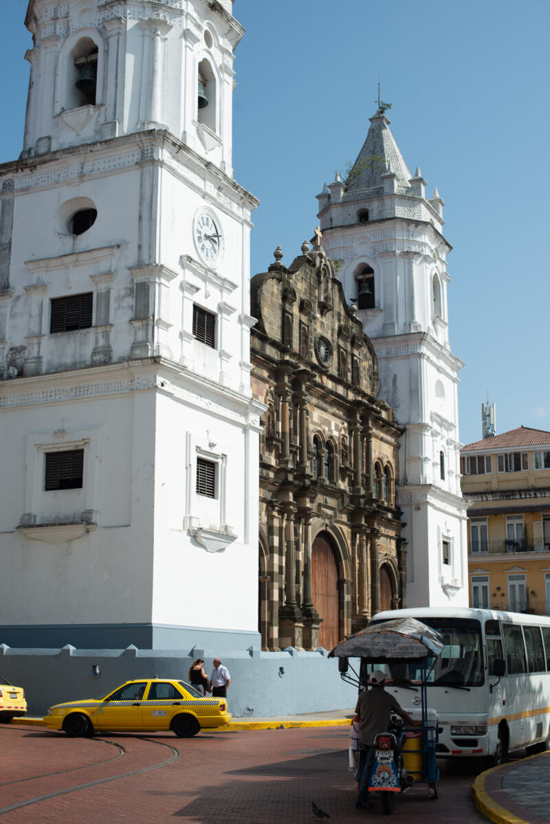 White bell tower with mother-of-pearl inlay and weathered stone central facade of the Catedral Metropolitana, the tower's clock and arched openings visible against a blue sky