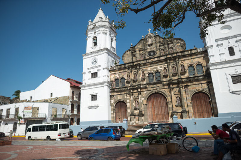 Catedral Basílica Santa María la Antigua seen from Plaza de la Independencia, its weathered stone central block flanked by two white bell towers, vehicles and a street vendor in foreground