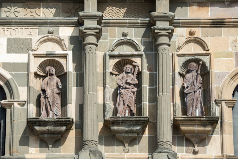 Three weathered wooden statues of robed saints in arched stone niches on the cathedral facade, each framed by pilasters and decorative stone carving