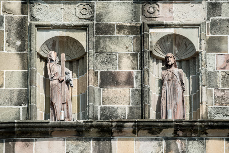 Two weathered wooden carved statues of robed saints in shell-backed stone niches on the cathedral facade, separated by fluted pilasters