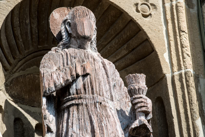 Weathered wooden statue of a saint holding a book or model church, seen from behind in a shell-backed niche on the cathedral facade