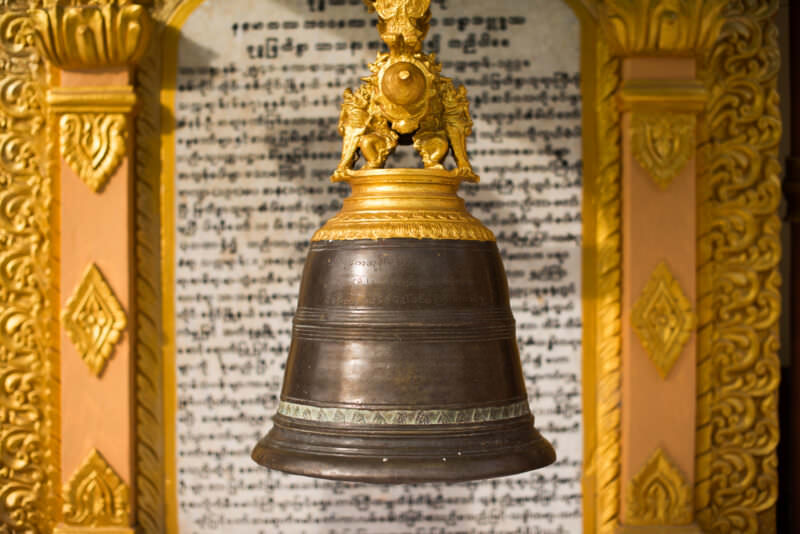 Bronze devotional bell with gilded mount hanging before a marble Burmese inscription board at U Min Thonze Pagoda
