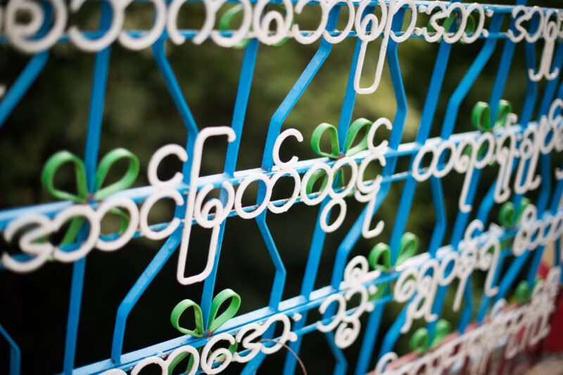 Curved Burmese script in white iron welded into blue metal fence bars at U Min Thonze Pagoda