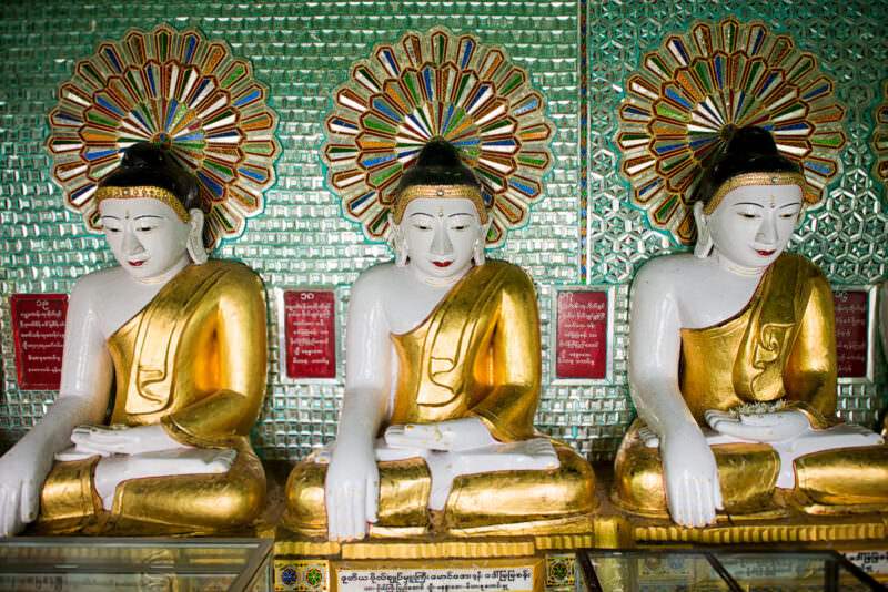 Three white-faced gilded Buddha images with colourful glass-mosaic halos and donor plaques at U Min Thonze Pagoda