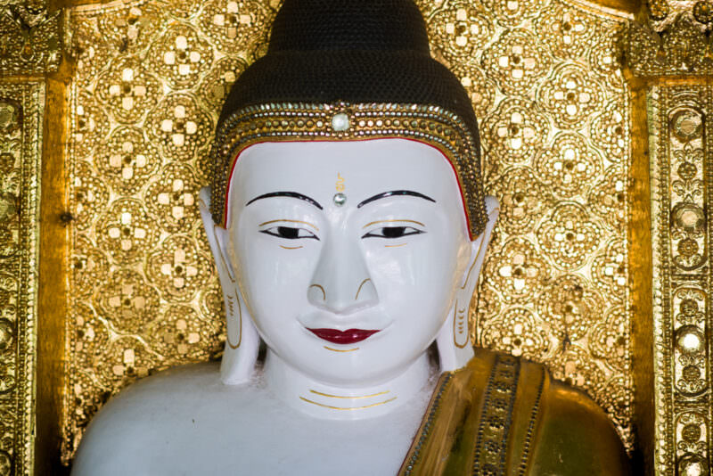 White-faced Buddha with gilded headband against gold floral glass-mosaic wall inside U Min Thonze Pagoda