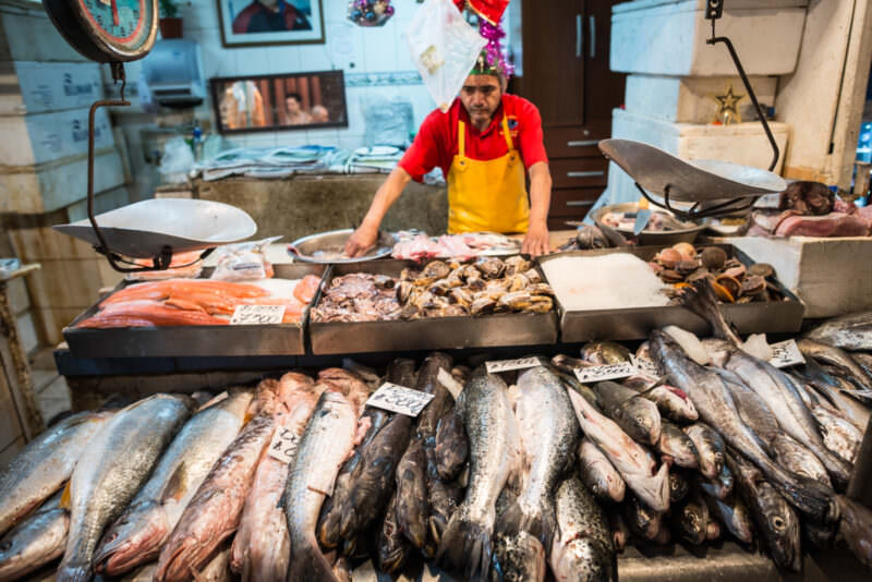 Vendor in yellow apron behind display of whole fish, salmon fillets, and shellfish with hanging scales at Mercado Central