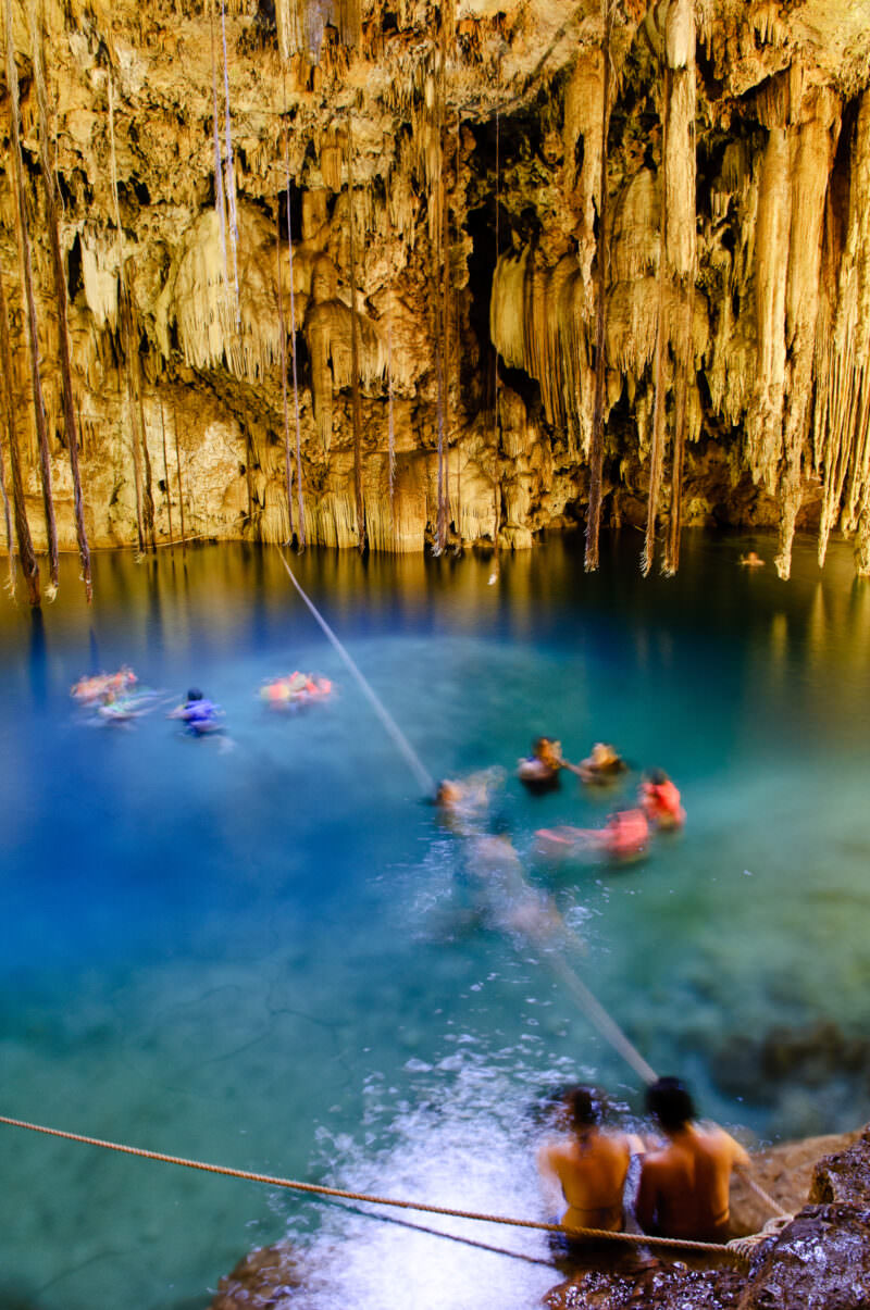 Swimmers in life jackets floating beneath dense stalactite formations at Cenote X'keken, lit by warm artificial lighting in the underground chamber