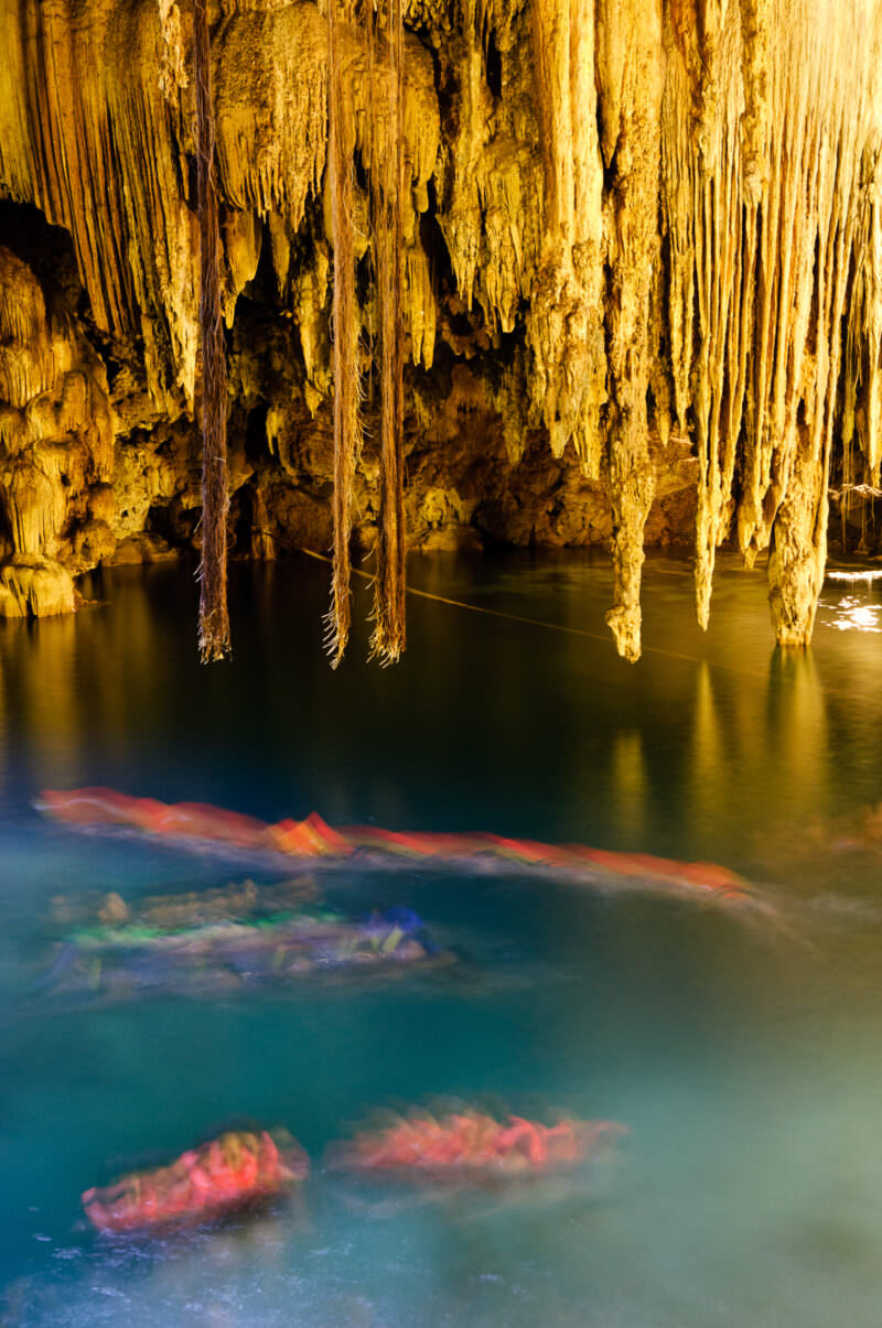 Life-jacketed swimmers floating in the illuminated turquoise waters of Cenote X'keken beneath long golden stalactites descending into the pool