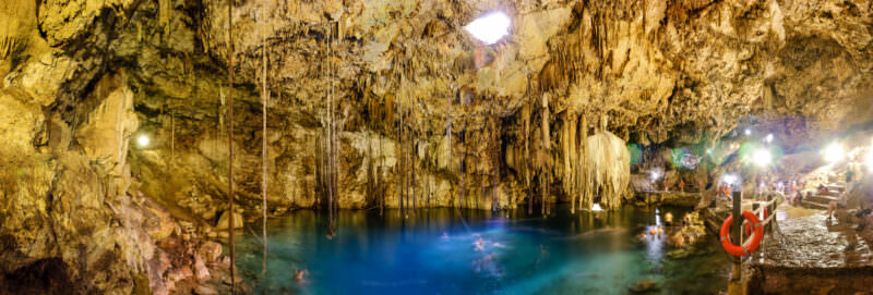 Wide panoramic view of Cenote Xkeken showing the full limestone cavern chamber with stalactites, turquoise waters, and natural light entering from the ceiling opening