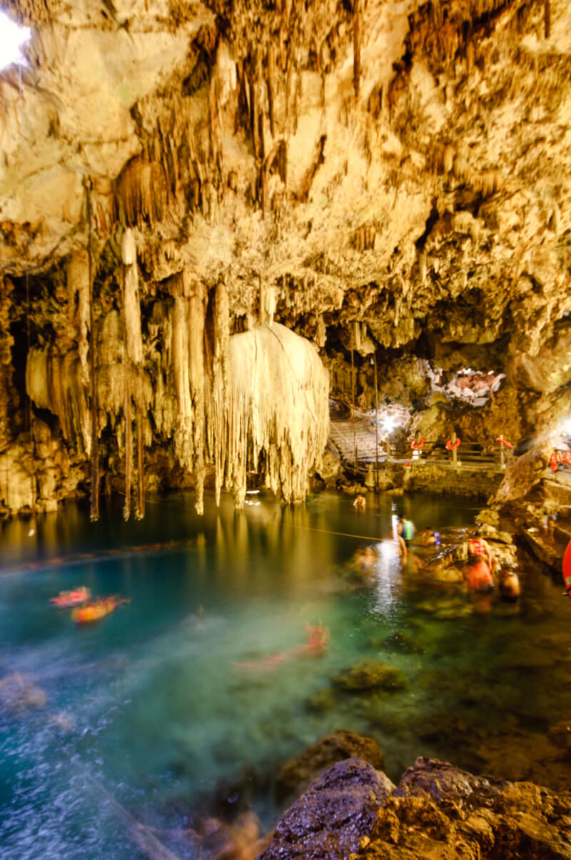 Cenote X'keken's cavern interior with swimmers near a stone stairway, surrounded by illuminated stalactite formations and limestone rock walls