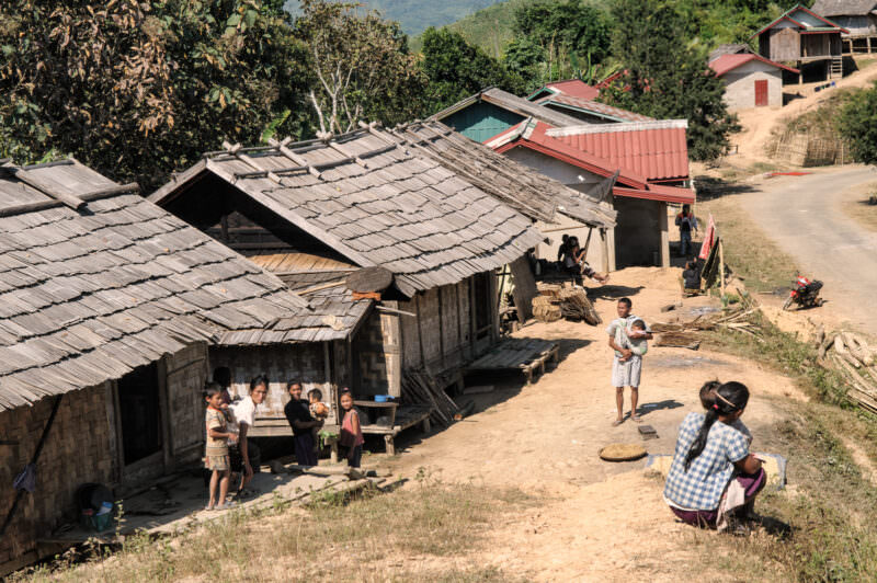 Wooden shingle-roofed stilt houses along a dirt road, villagers gathered outside, forested mountains in background