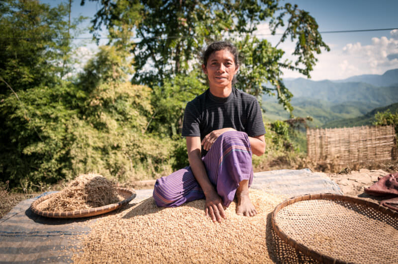 Woman in purple skirt sitting on a tarp of drying rice grains, bamboo winnowing trays beside her, mountains behind