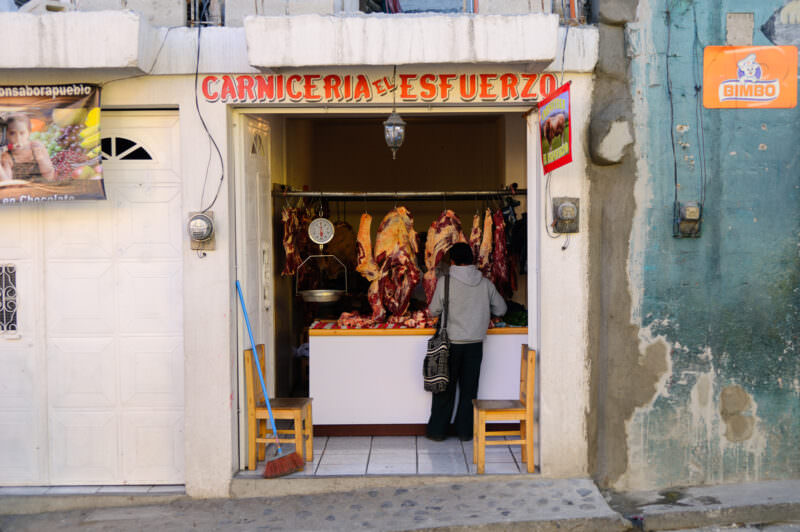 Small storefront butcher shop Carniceria El Esfuerzo with hanging meats visible through open doorway, customer inside
