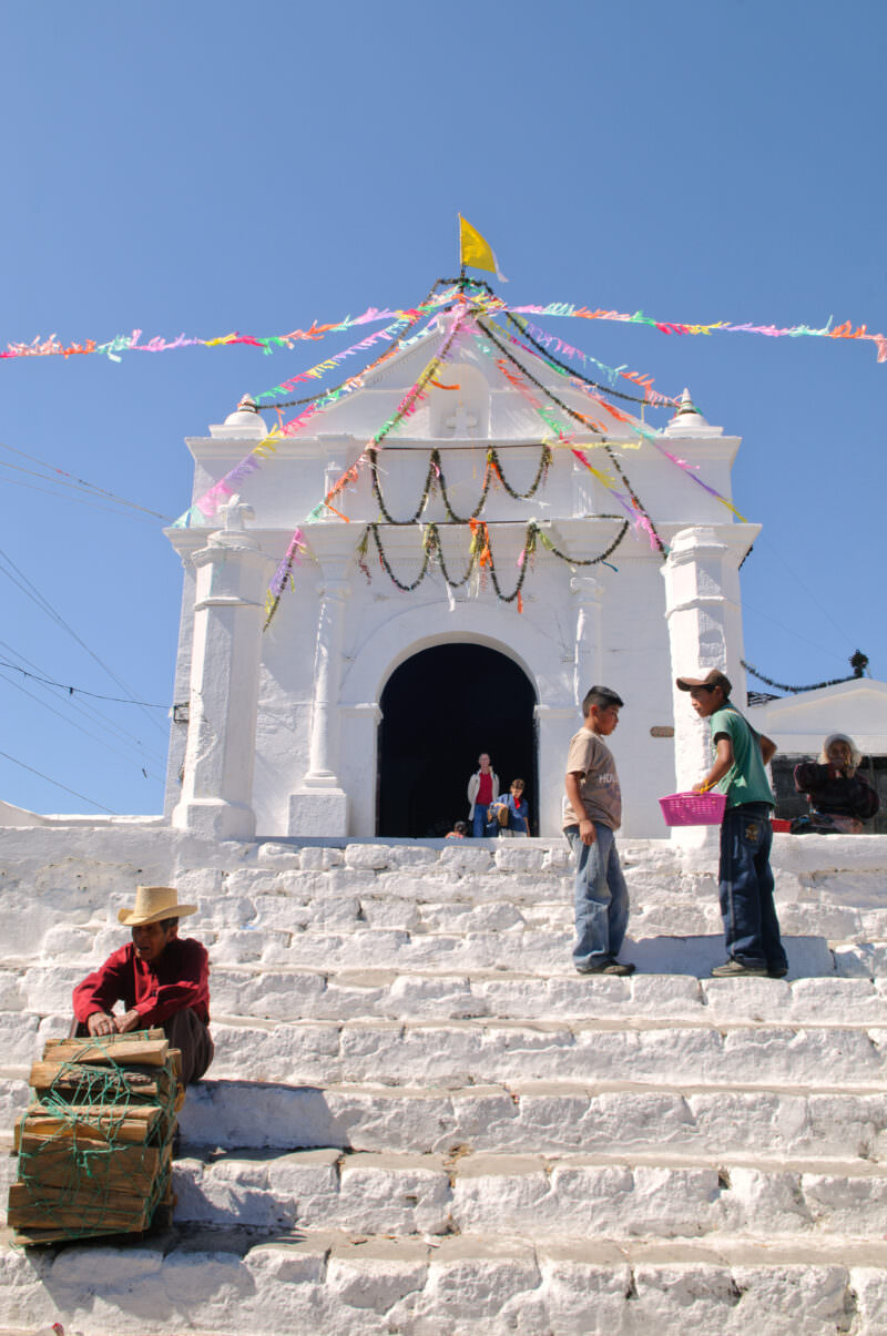 Capilla del Calvario chapel with white stepped facade, colorful pennant banners radiating from peak, firewood vendor at base