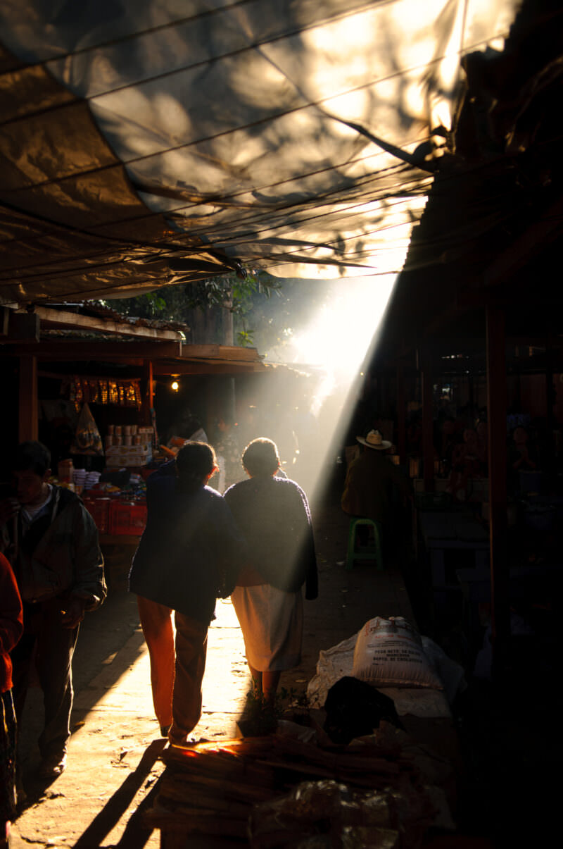 Bright sunbeam streaming through opening in dark covered market roof, silhouetted shoppers walking below