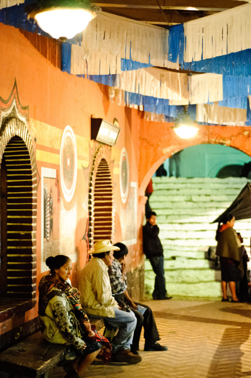 Three people seated on bench under orange arched colonnade with blue and white paper decorations, church steps visible beyond