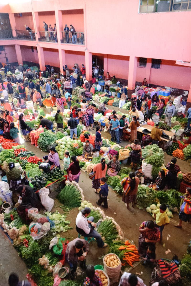 Aerial view of indoor produce market in pink gymnasium courtyard, vendors and shoppers amid piles of fresh vegetables
