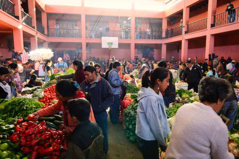 Crowded indoor produce market courtyard surrounded by pink two-story building, vendors displaying vegetables and greens below